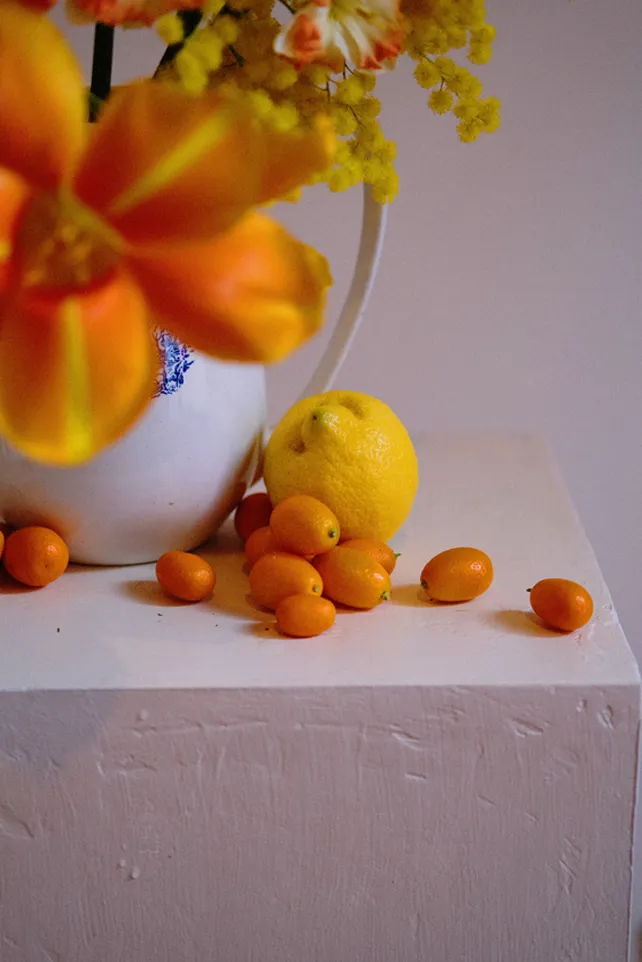 sur un caisson plâtré et brossé de couleur rose se trouvent des fruits de couleur orange et jaune avec un vase contenant des fleurs orange et jaune