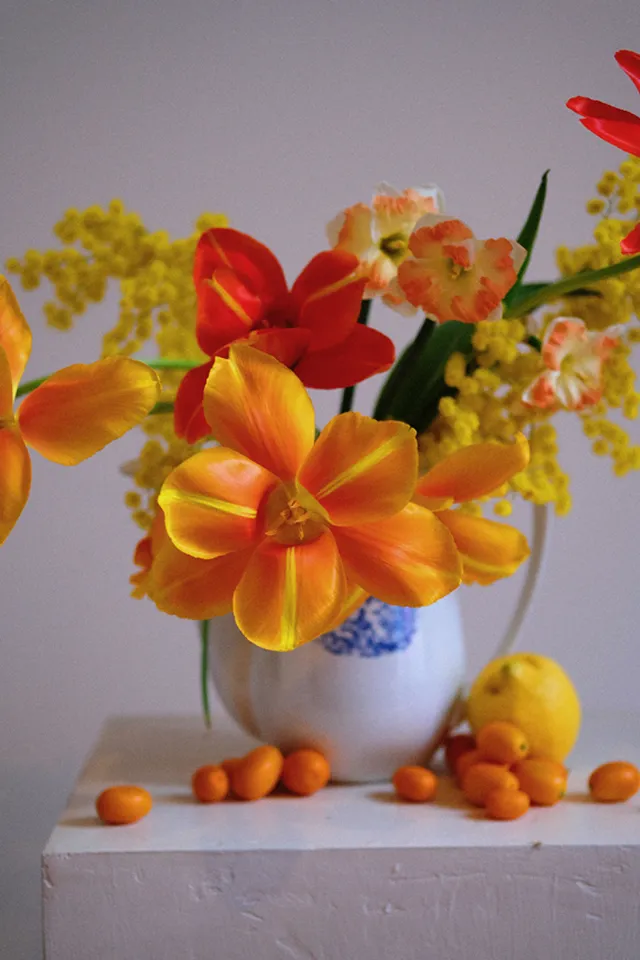 une composition florale de fleurs orange et rouge dans un vase blanc, posé sur un caisson en plâtre rose, comprenant des fruits de couleur orange et jaune