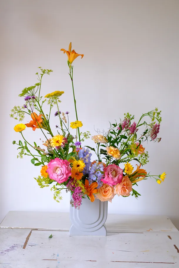 dans un joli vase en forme d’U de couleur blanche se trouvent différentes fleurs des couleurs rose pâle, rose vif, jaune, orange, violet et des feuilles vertes
