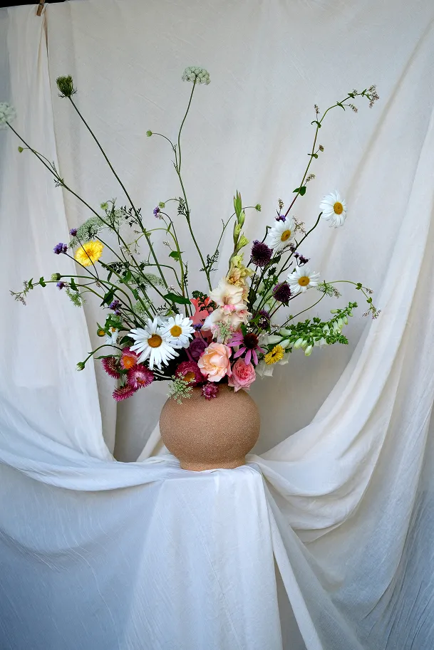 dans un vase éloigné posé sur un fond de nappe se trouve un bouquet de fleurs comprenant des fleurs de couleurs blanches, rose pâle, rose vif, ainsi que des tiges et des feuilles vertes