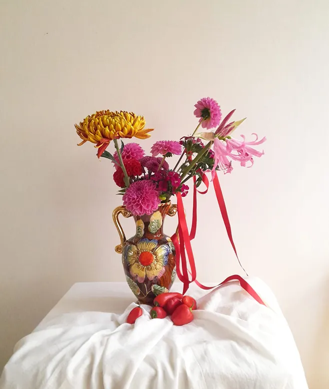 sur une table recouverte d’une nappe blanche se trouvent des piments et poivrons, ainsi qu’un vase avec un décor de fleurs, et dans ce vase se trouvent des fleurs roses et jaunes
