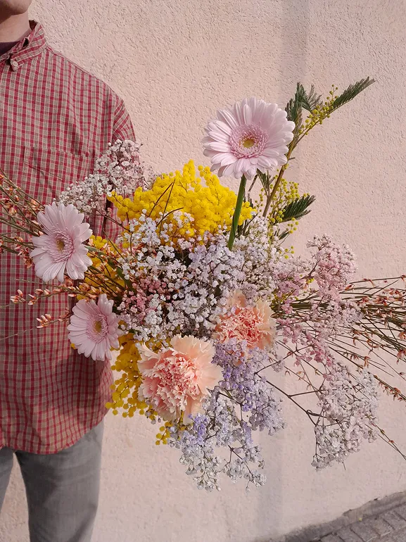 un homme tient dans sa main, de face, un bouquet de fleurs composé des couleurs rose, blanc et jaune