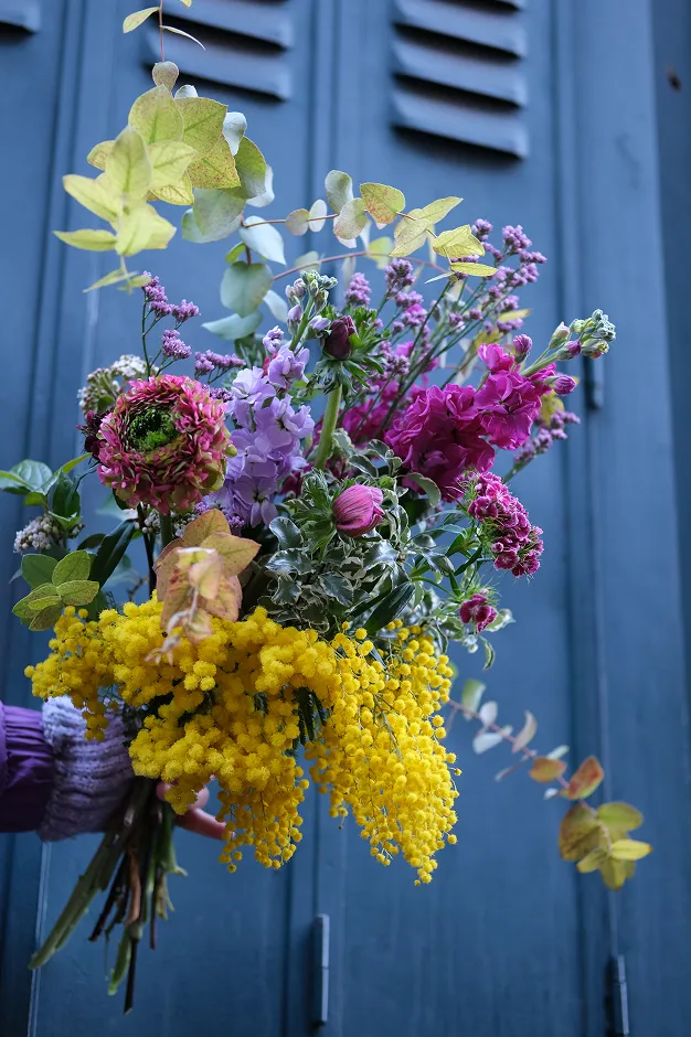 sur fond bleu une main tient un bouquet de fleurs avec les couleurs jaune, rose, violet et blanc