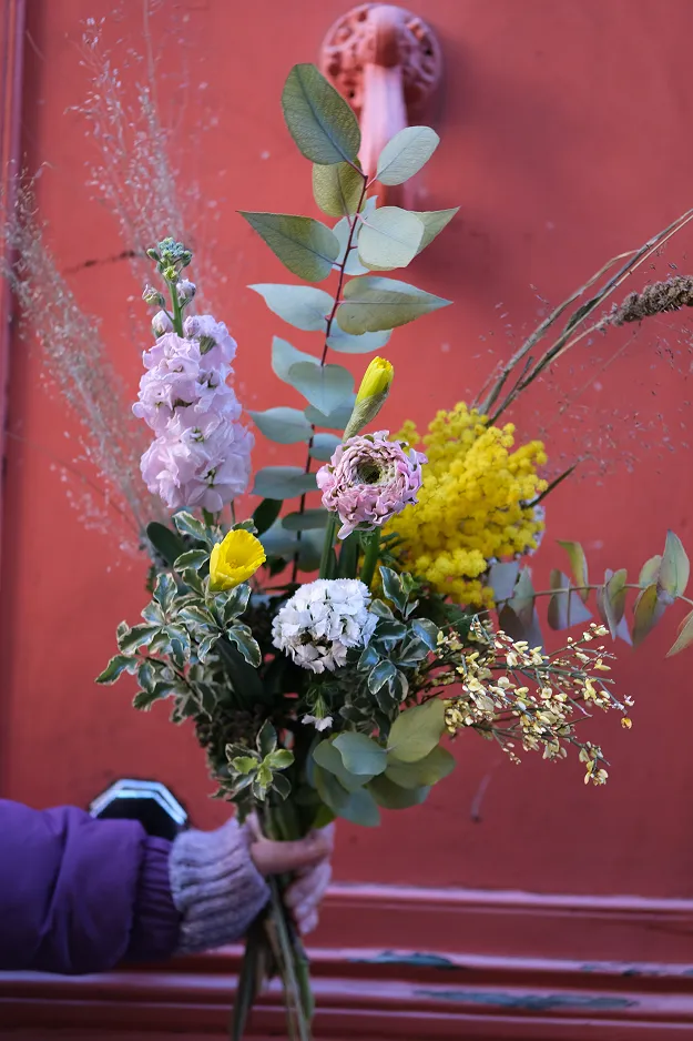 sur un fond rouge une main tient un petit bouquet avec des feuilles vertes et des fleurs de couleur rose, jaune et blanche