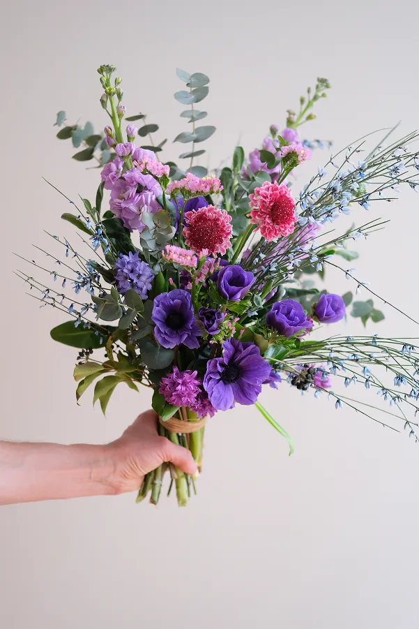 dans une main se tient un bouquet composé de feuilles vertes avec des fleurs de couleur violette, rose vif et rose clair