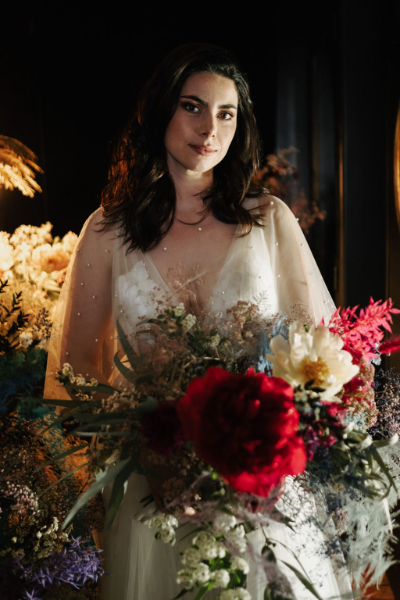 une femme vêtue d’une robe de mariée dans un couloir, rempli de fleurs de toutes les couleurs, tenant un bouquet de fleurs composées des couleurs rouge, blanc, rose et vert