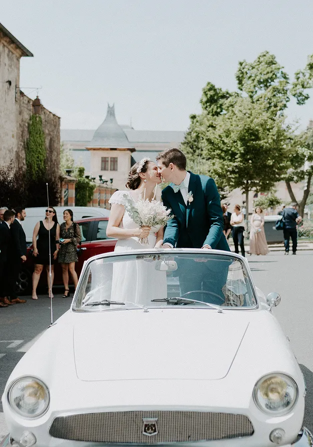 dans un village, avec un arrière-plan, un bâtiment, des arbres et des personnes discutant lors d’un mariage, et en premier plan, dans une voiture blanche décapotable, debout, se trouvent le futur mari et la future mariée s’embrassant ; la mariée tient dans ses mains un bouquet de fleurs blanches et est vêtue d’une robe de mariée blanche