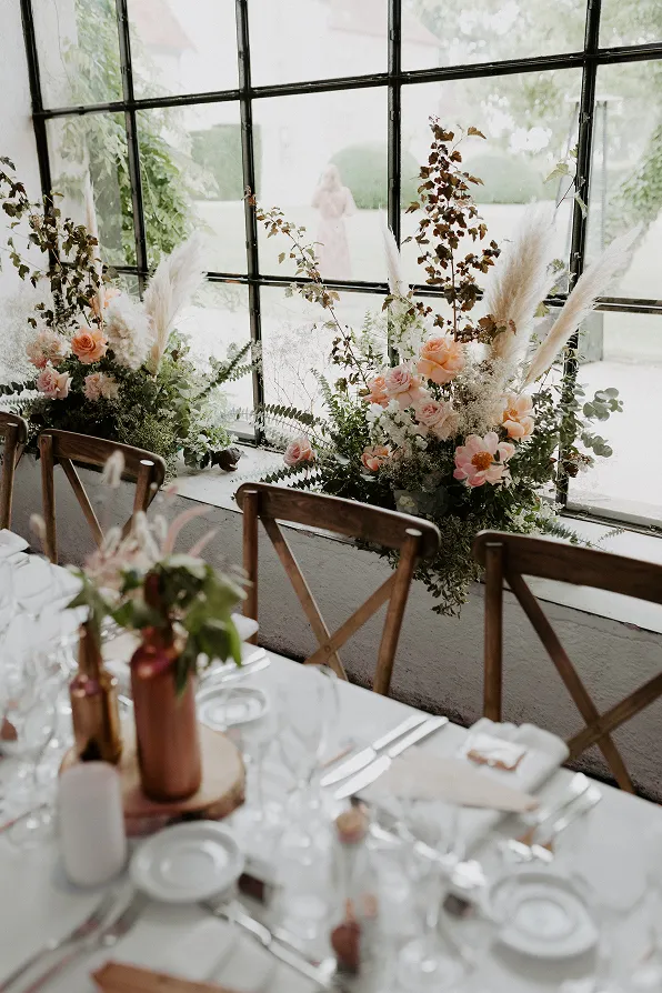 une table avec des couverts et, en arrière-plan, des fleurs lors d’un mariage, composées des couleurs blanche et rose sur un fond vitré de type ancien
