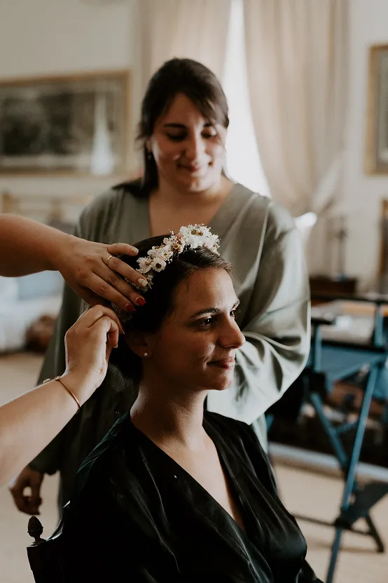 une femme assise et une femme debout en arrière ; la femme assise est en train de se faire coiffer, et on lui installe un serre-tête de fleurs blanc