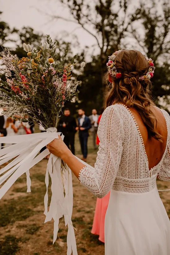 une femme vêtue d’une robe de mariée de dos tient dans sa main un bouquet de fleurs, enrobé de ruban blanc et composé des couleurs jaune, rose et rouge, accompagné de feuilles vertes