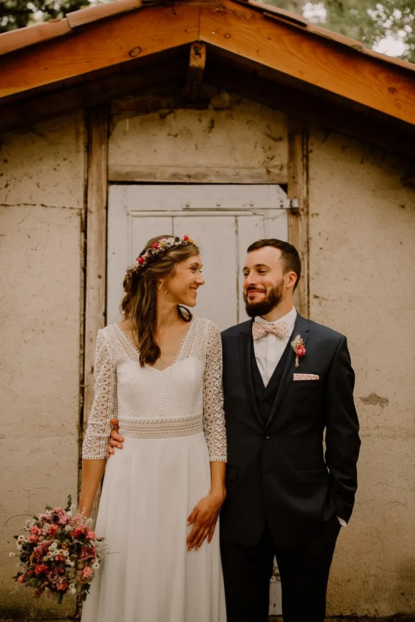 un couple de mariés avec une femme vêtue d’une robe de mariée, tenant dans sa main droite un bouquet de fleurs roses et blanches, et un homme en costume bleu marine foncé avec un col blanc et un nœud papillon rose pâle