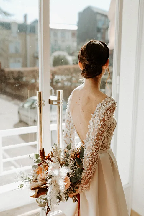 une femme vêtue d’une robe de mariée, se tenant de dos et tenant dans ses mains, à l’arrière de son dos, un bouquet de fleurs composé des couleurs blanc et rouge avec des feuilles vertes