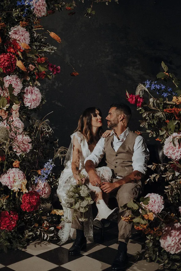 en premier plan se trouve une grande densité de fleurs de diverses couleurs, comprenant du rose, du rouge, de l’orange, du bleu et du vert, et derrière se trouve un couple de mariés se regardant avec affection