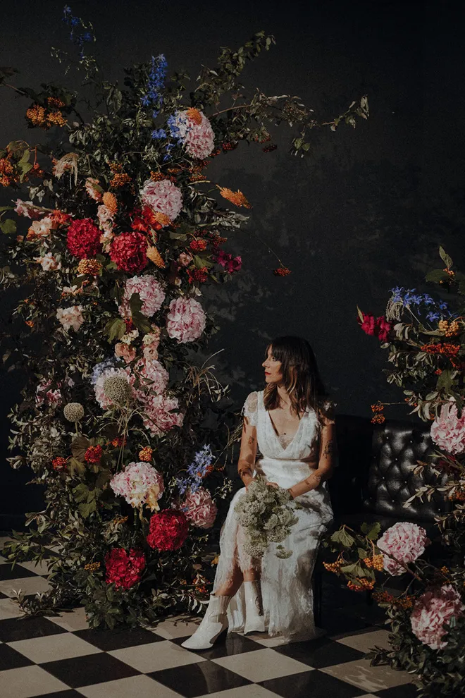 dans un décor rempli de fleurs avec une grande densité et de couleurs diverses se trouve une femme vêtue d’une robe de mariée, tenant un bouquet de fleurs blanches