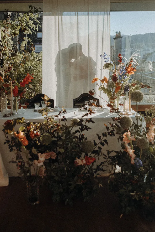 dans un décor rempli de fleurs derrière un voile blanc se trouve un couple de mariés s’embrassant