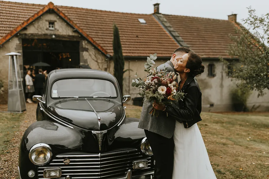 à côté d’une voiture noire et ancienne se trouve un couple de mariés
