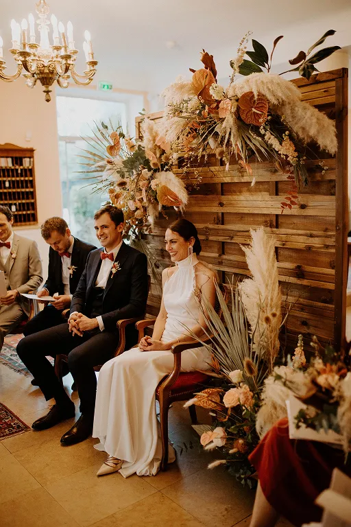 un couple de mariés, assis chacun sur une chaise, entouré d’un décor de fleurs