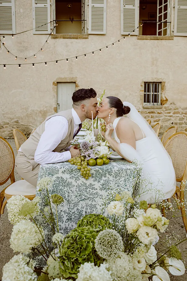 lors d’un mariage, les mariés s’embrassent autour d’une table avec une composition florale, un verre blanc et bleu