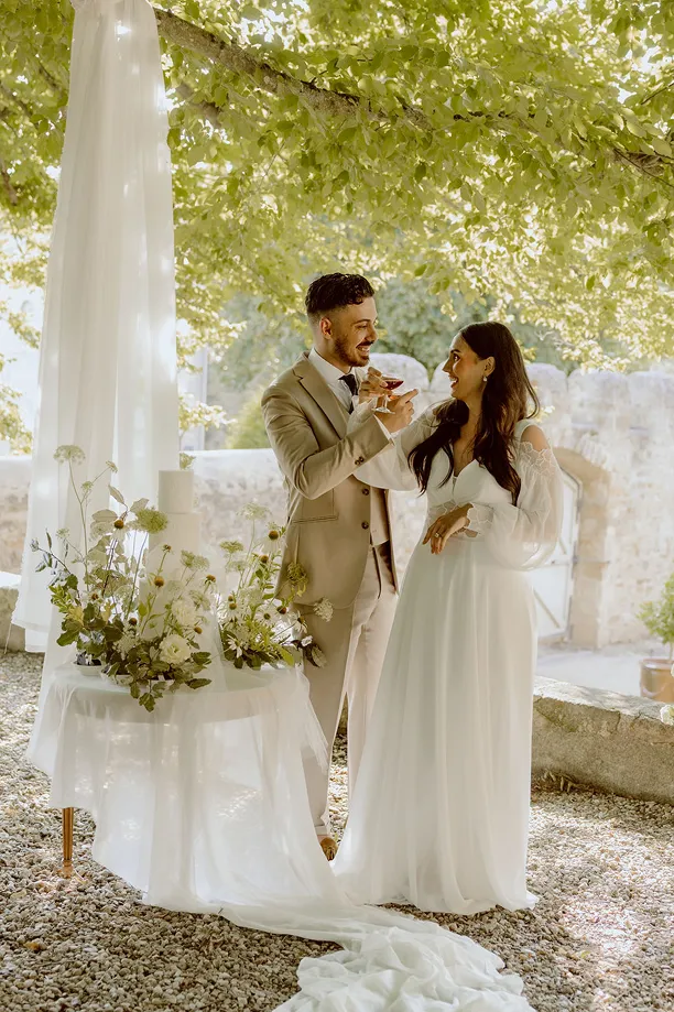 un couple de mariés lors de leur mariage tiennent chacun une coupe de cocktails et se croisent les bras pour boire leur verre
