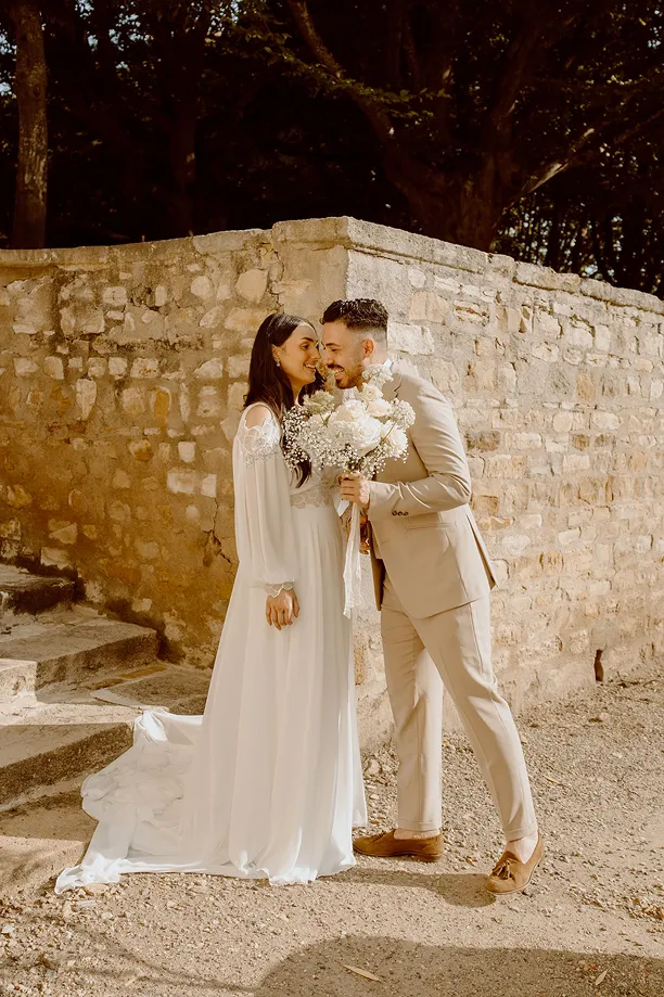 en extérieur, photo d’un couple marié ; l’homme tient dans sa main des fleurs blanches et s’approche de la mariée pour l’embrasser